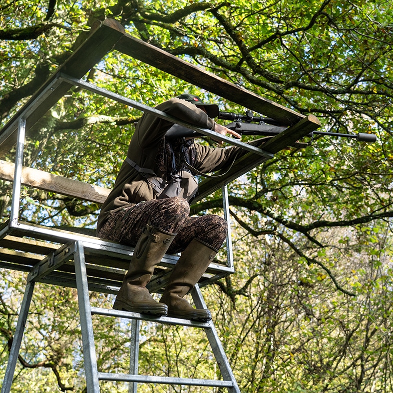 A deer stalking aiming their rifle whilst sitting in a high seat